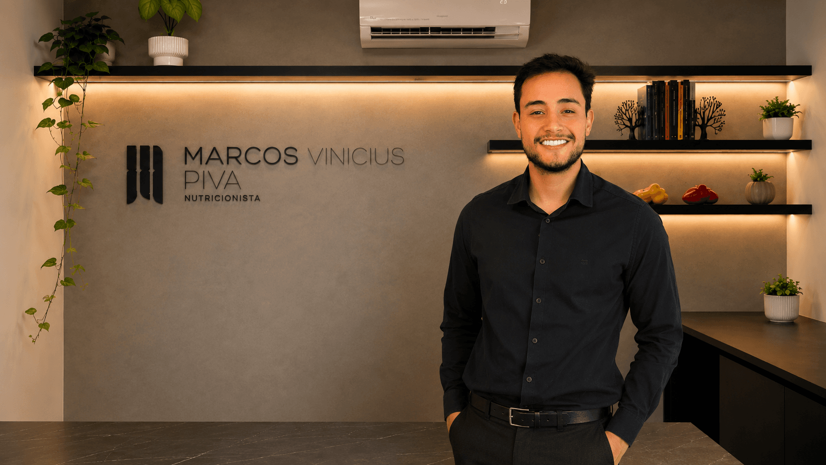 Marcos Vinicius Flores Piva sorrindo em seu consultório em Bonito - MS, com a parede de cimento queimado e as prateleiras iluminadas ao fundo